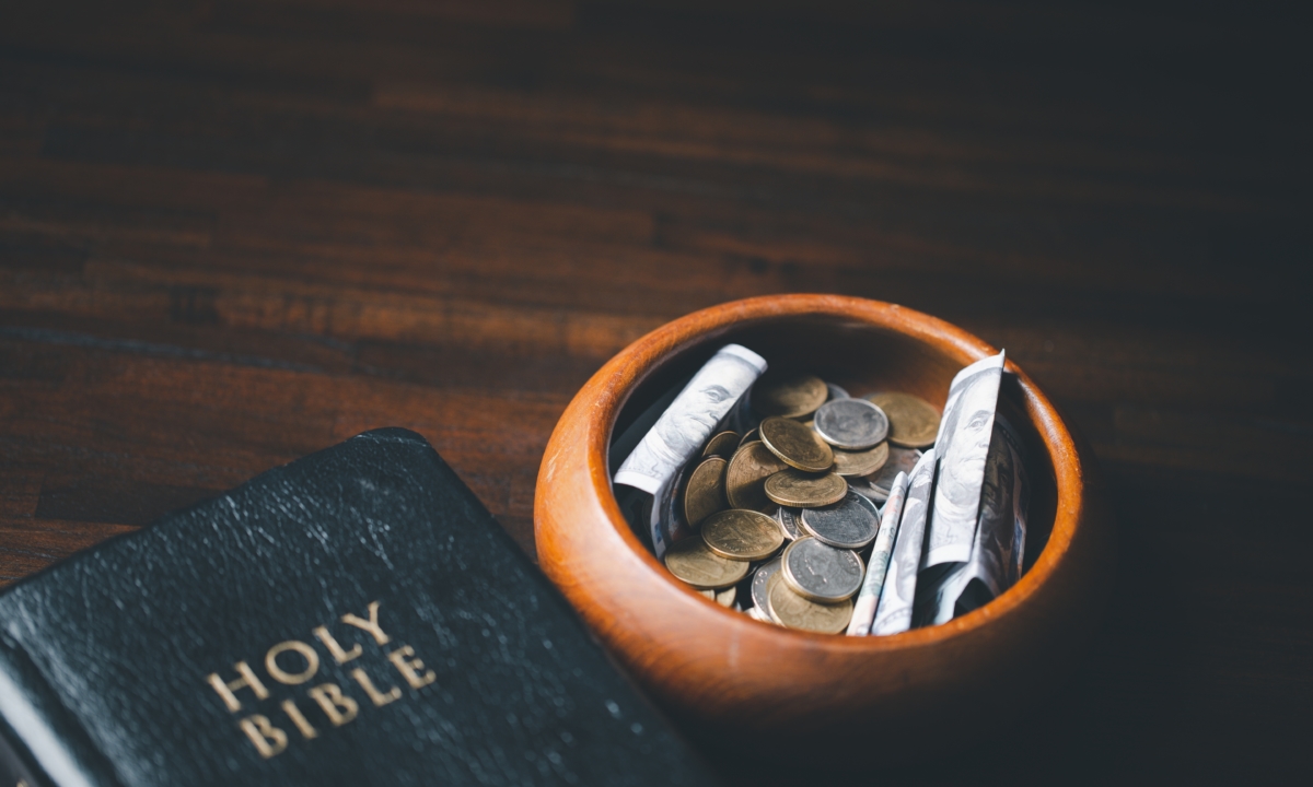 Dollars and coins inside a bowl next to a Bible on a table